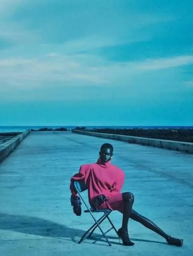 Model in a bright pink outfit seated on a folding chair on a concrete pier with blue sky and water behind