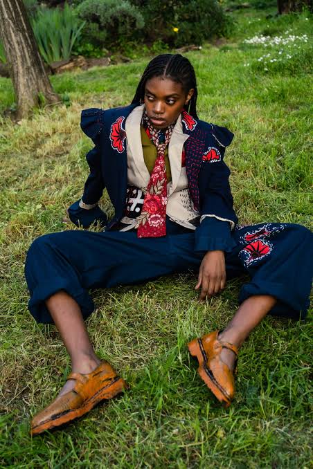 Model seated on grass in a navy embroidered jacket and cropped trousers